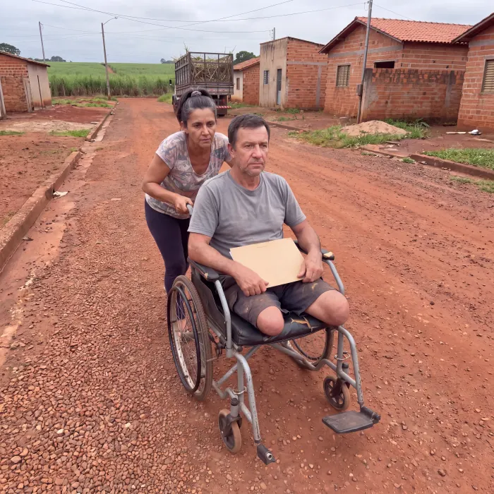 Uma mulher empurra a cadeira de rodas de Valdir por uma estrada de terra entre as casas do povoado